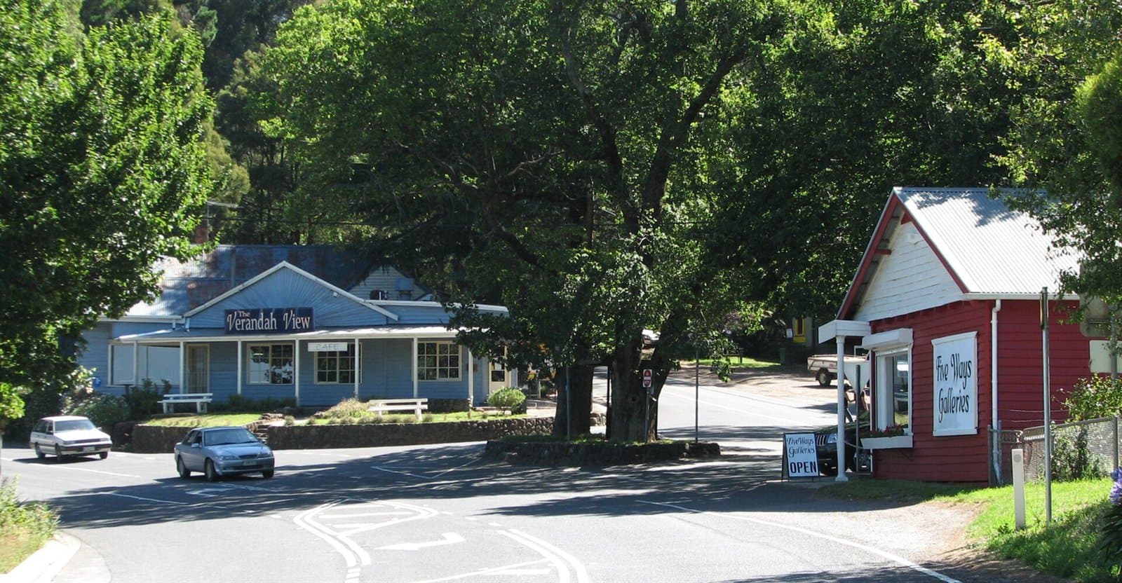 A road junction under large oak trees, with a weatherboard building on each side