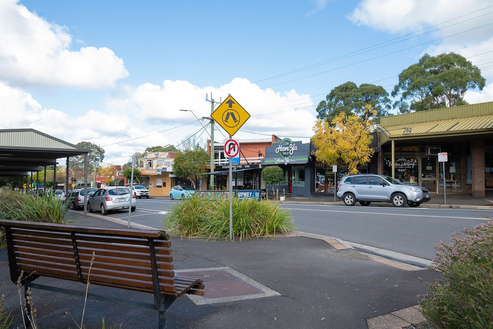 Shops and parked cars along the main street of Upwey, with a bench in the foreground
